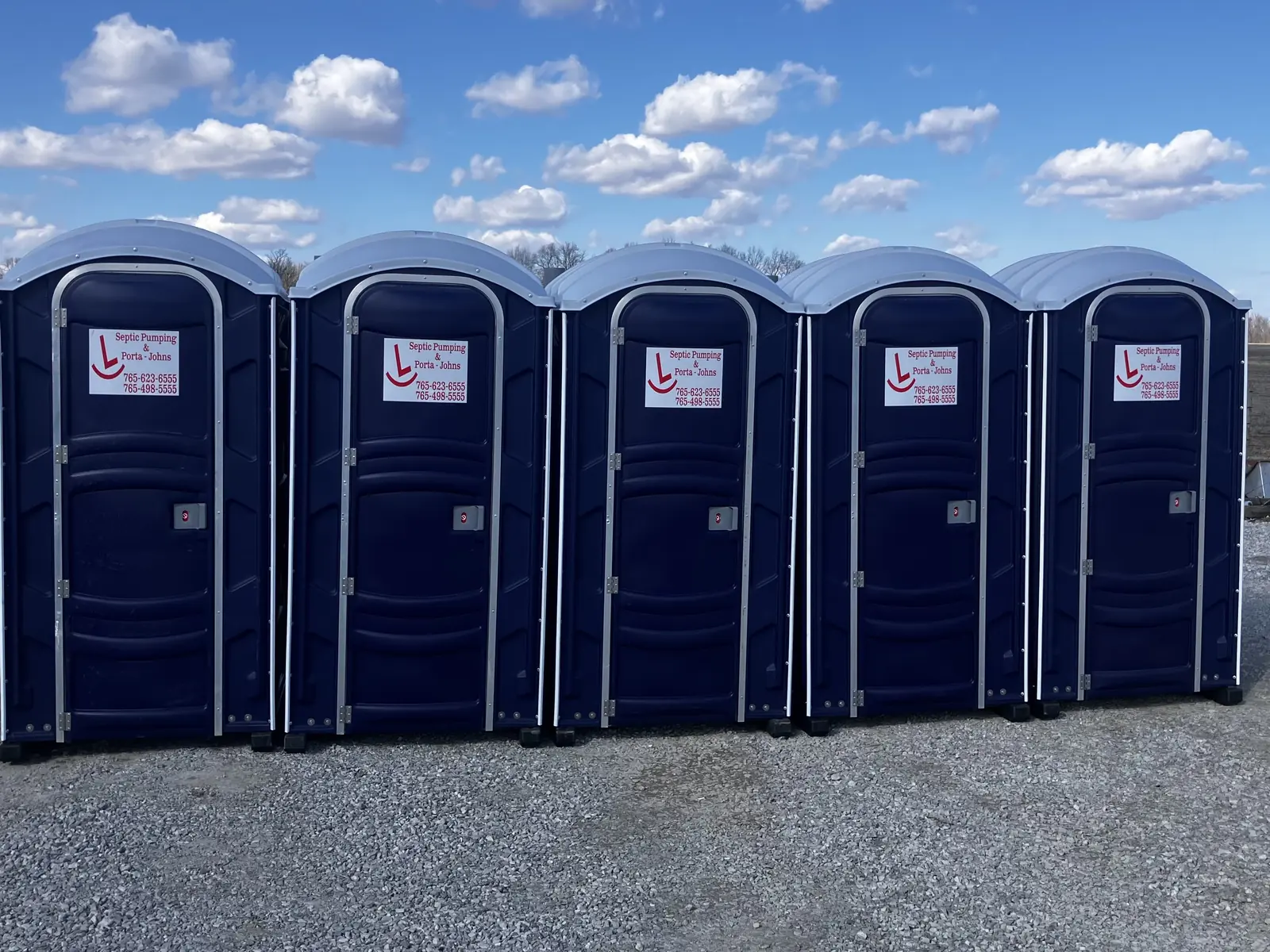Five blue Rockin L portable restrooms lined up on a gravel lot under a clear Indiana sky, ready for delivery