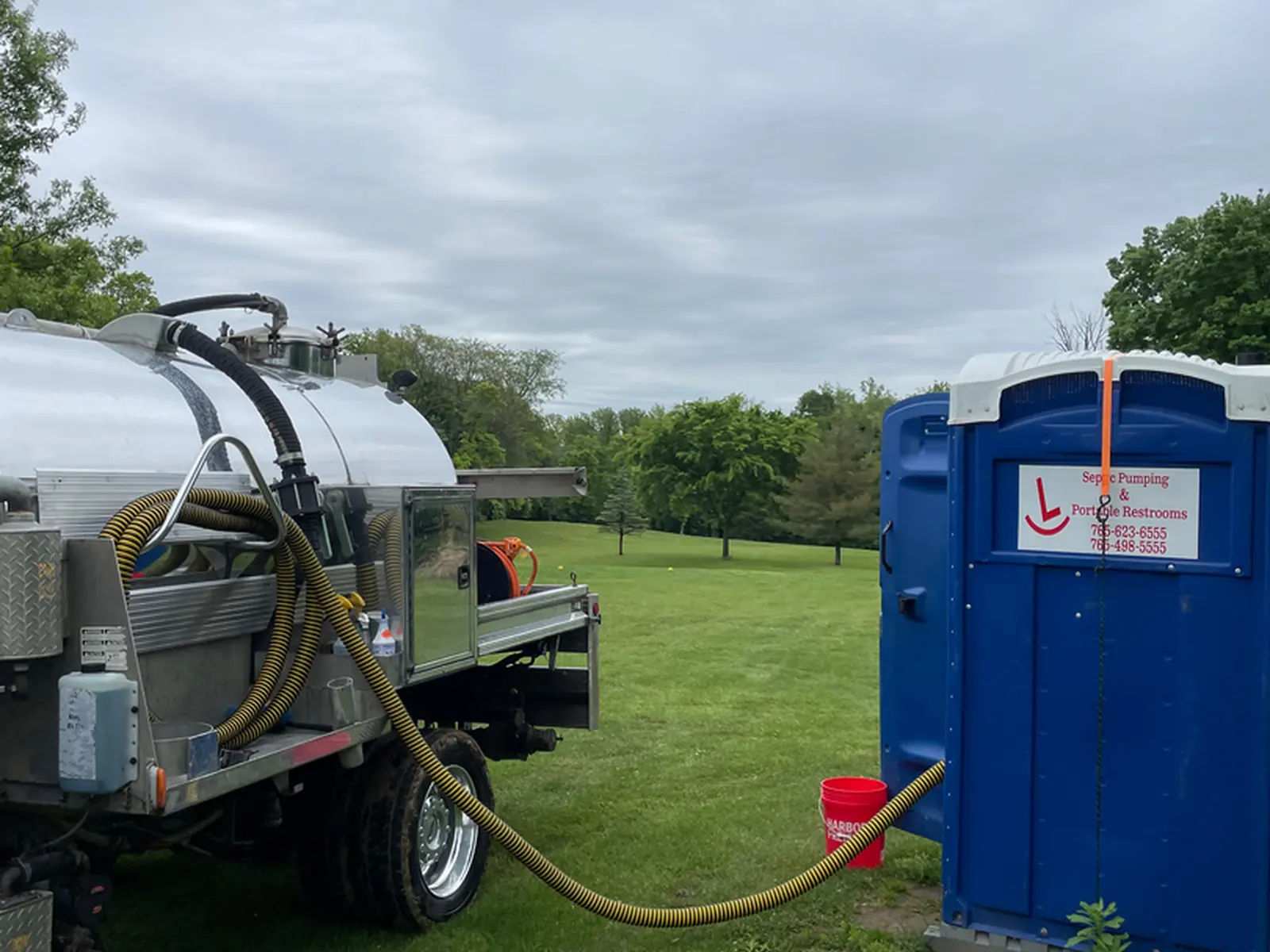 Rockin L vacuum tank truck with a yellow suction hose running across a rural Indiana lawn to an open septic tank