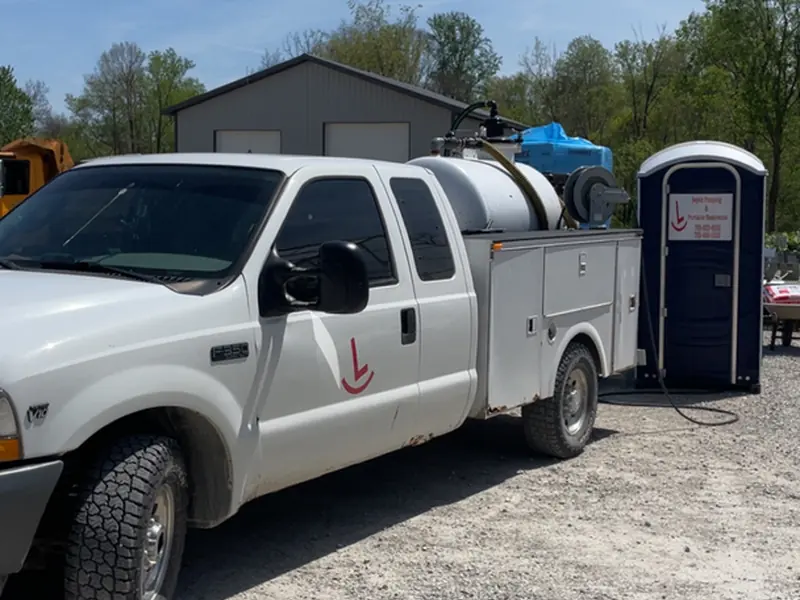A white Rockin L service truck with a mounted tank and pump equipment parked in the yard beside a portable restroom