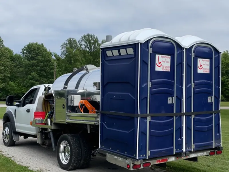 A flatbed truck hauling two strapped Rockin L portable restrooms out of the yard