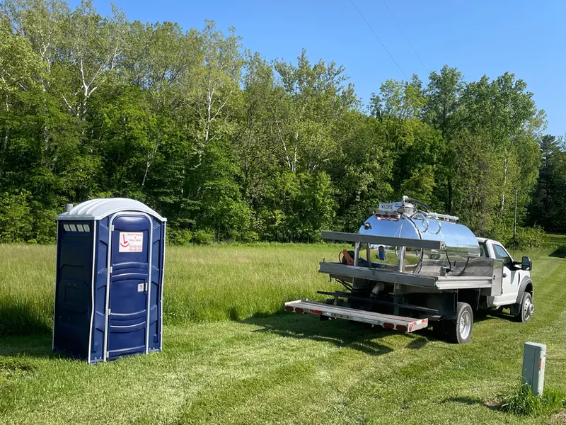 A Rockin L portable restroom and a flatbed truck with a stainless vacuum tank parked in a mowed field in rural west-central Indiana
