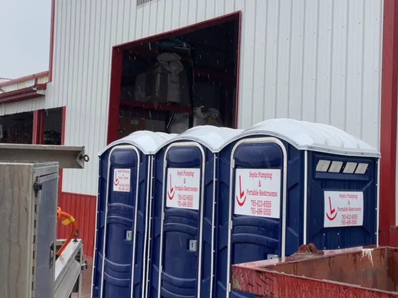 Two Rockin L portable restrooms being serviced on a wet gravel lot beside a metal building