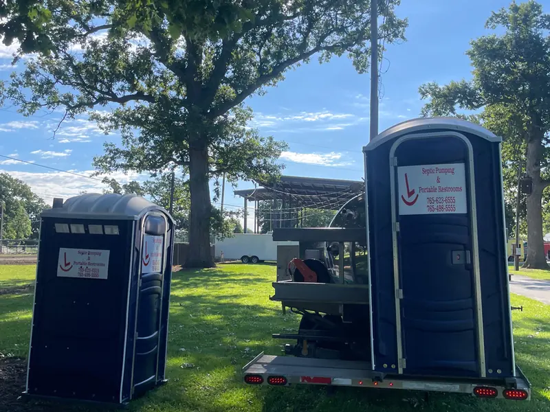 Rockin L portable restrooms on a delivery trailer parked under mature shade trees at an event ground